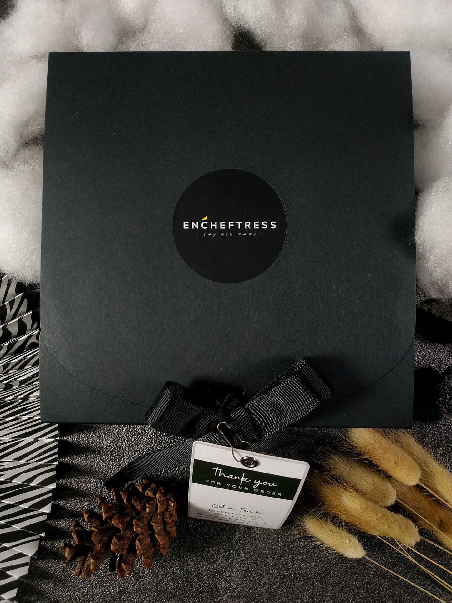 A closed black gift box with a circular logo, a black ribbon, and a "Thank you for your order" tag, surrounded by fluffy white material, feathery plumes, a pinecone, and a striped pattern.