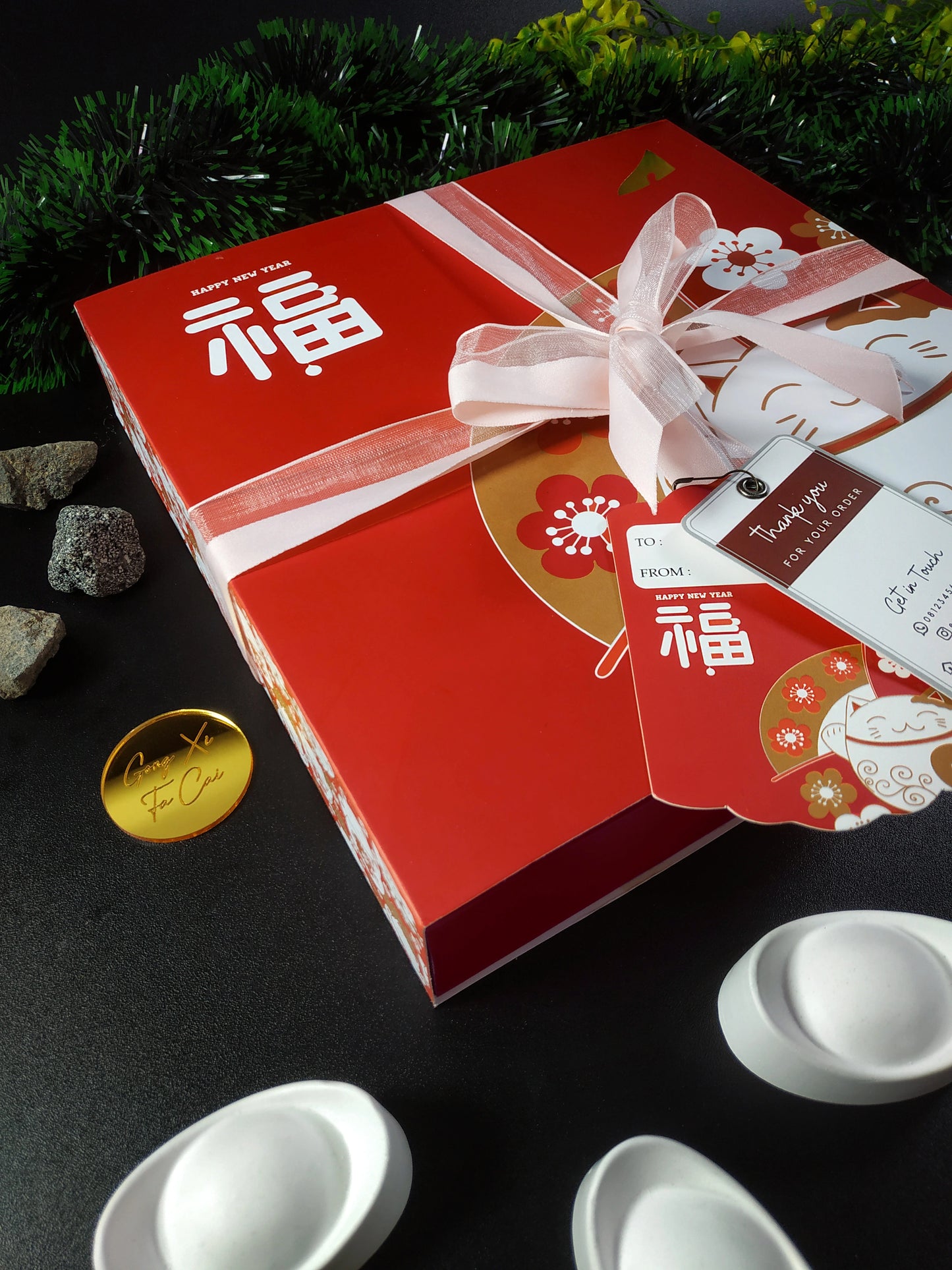 A red Lunar New Year gift box with gold lettering and a white and pink ribbon, featuring a "Thank you for your order" tag, a round gold coin, decorative stones, and a partial view of white packaging and green tinsel.