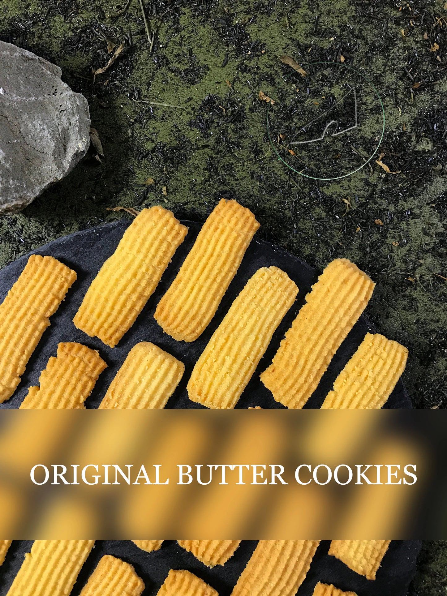 Several rectangular butter cookies with ridged tops are arranged on a dark, possibly slate, surface. A gray stone is visible in the upper left corner, and the background is a textured dark green. The text "ORIGINAL BUTTER COOKIES" is overlaid across the center of the image with a blurred effect.