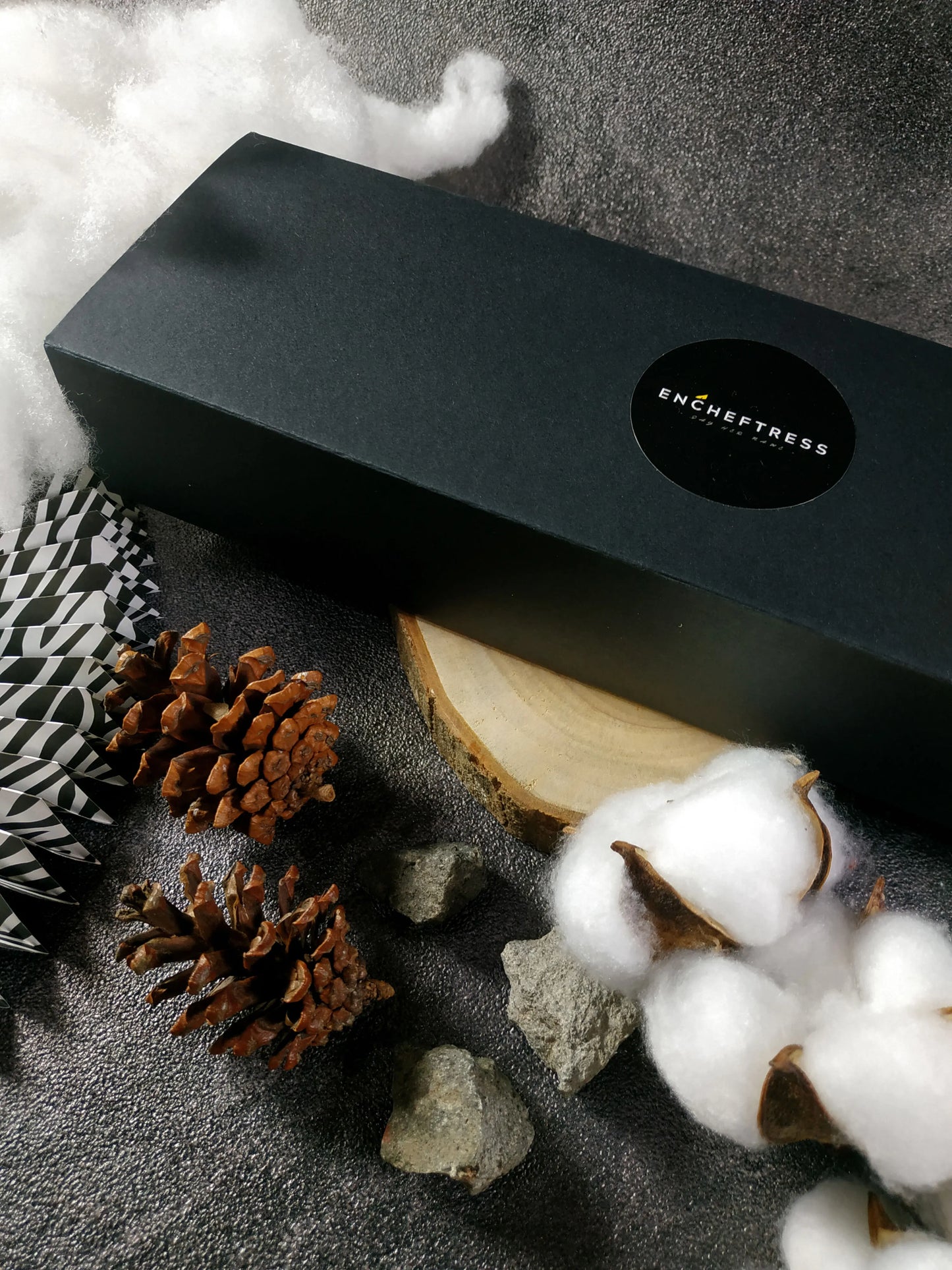 A closed black gift box from 'ENCHEFTRESS' styled with pinecones and cotton bolls. Contains 5 Choux au Craquelin.