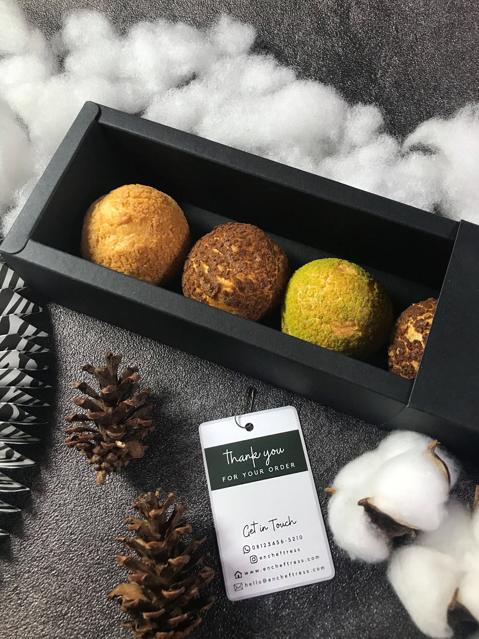Open black 'encheftress' box showing four colorful Choux au Craquelin pastries, styled with pinecones, cotton bolls, and a thank you tag. Box contains 5 pastries.
