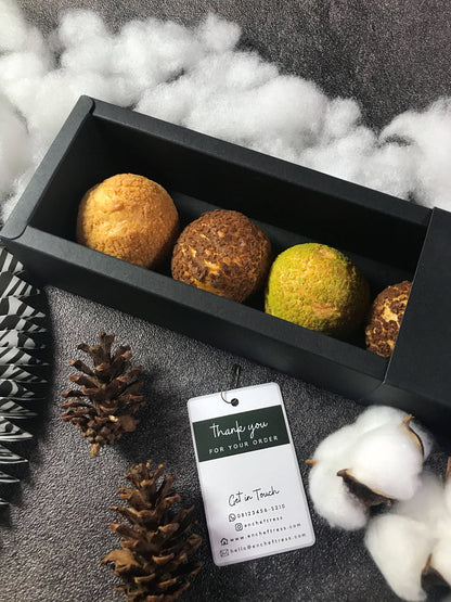 Open black 'encheftress' box showing four colorful Choux au Craquelin pastries, styled with pinecones, cotton bolls, and a thank you tag. Box contains 5 pastries.