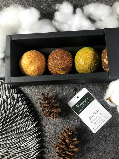 Top-down view of an open black box with four visible Choux au Craquelin pastries. Includes pinecones, cotton, patterned paper, and an 'encheftress' thank you tag. Box contains 5 pastries.