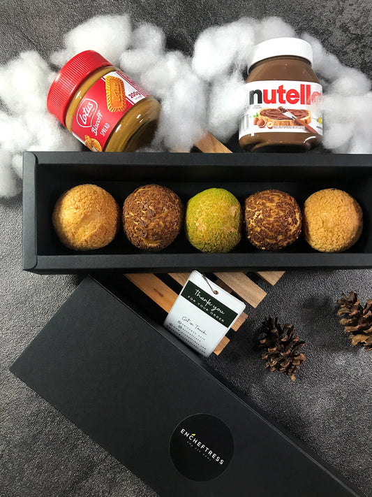 Five choux au craquelin pastries in a black box, with a jar of Lotus Biscoff spread and Nutella in the background, arranged on a textured surface with decorative cotton and pinecones.