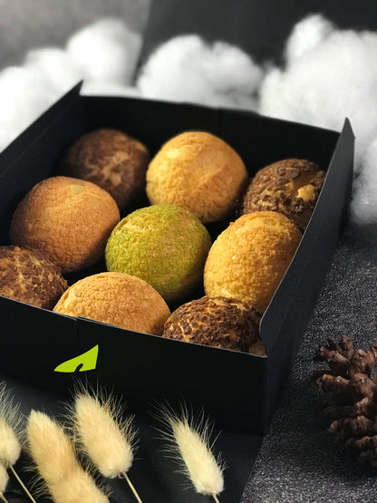 Nine colorful choux au craquelin pastries nestled in a black box, with fluffy white decorations, feathery plumes, and a pinecone surrounding it.
