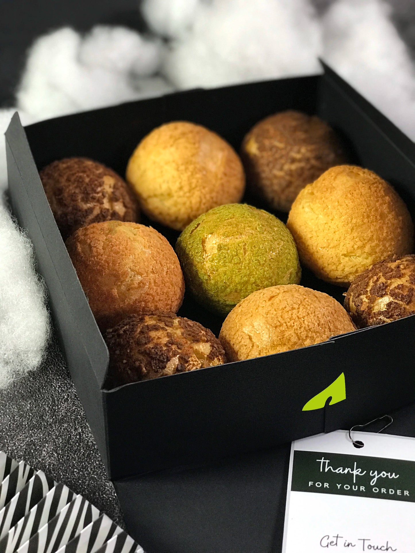 Nine colorful choux au craquelin pastries are neatly arranged in a black gift box, set against a backdrop of fluffy white material and a dark surface. A "Thank you for your order" tag is visible near the bottom.