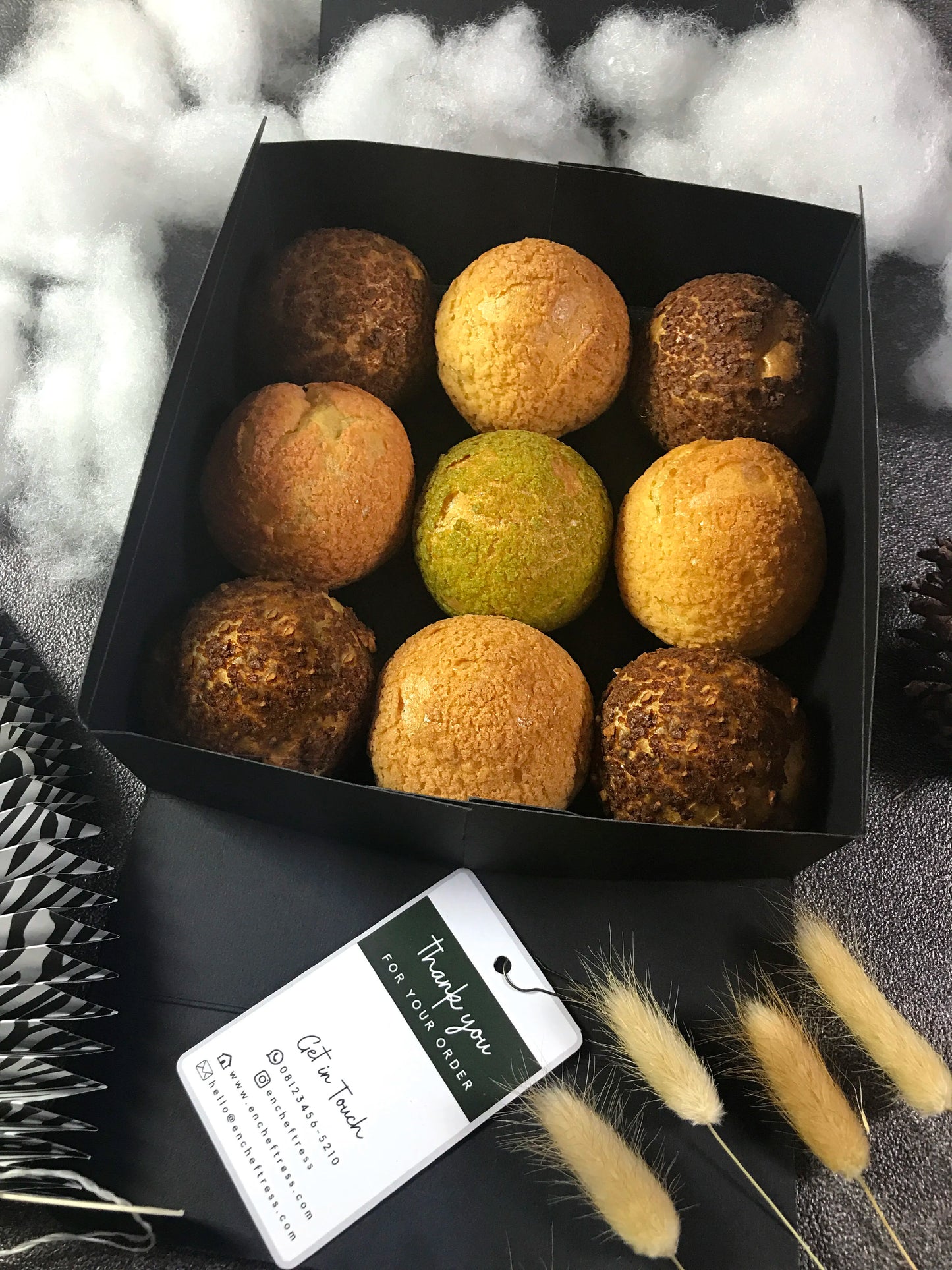 Nine colorful choux au craquelin pastries are arranged inside a black square box, accompanied by a "Thank you for your order" tag and surrounded by fluffy white material, feathery plumes, and a pinecone.