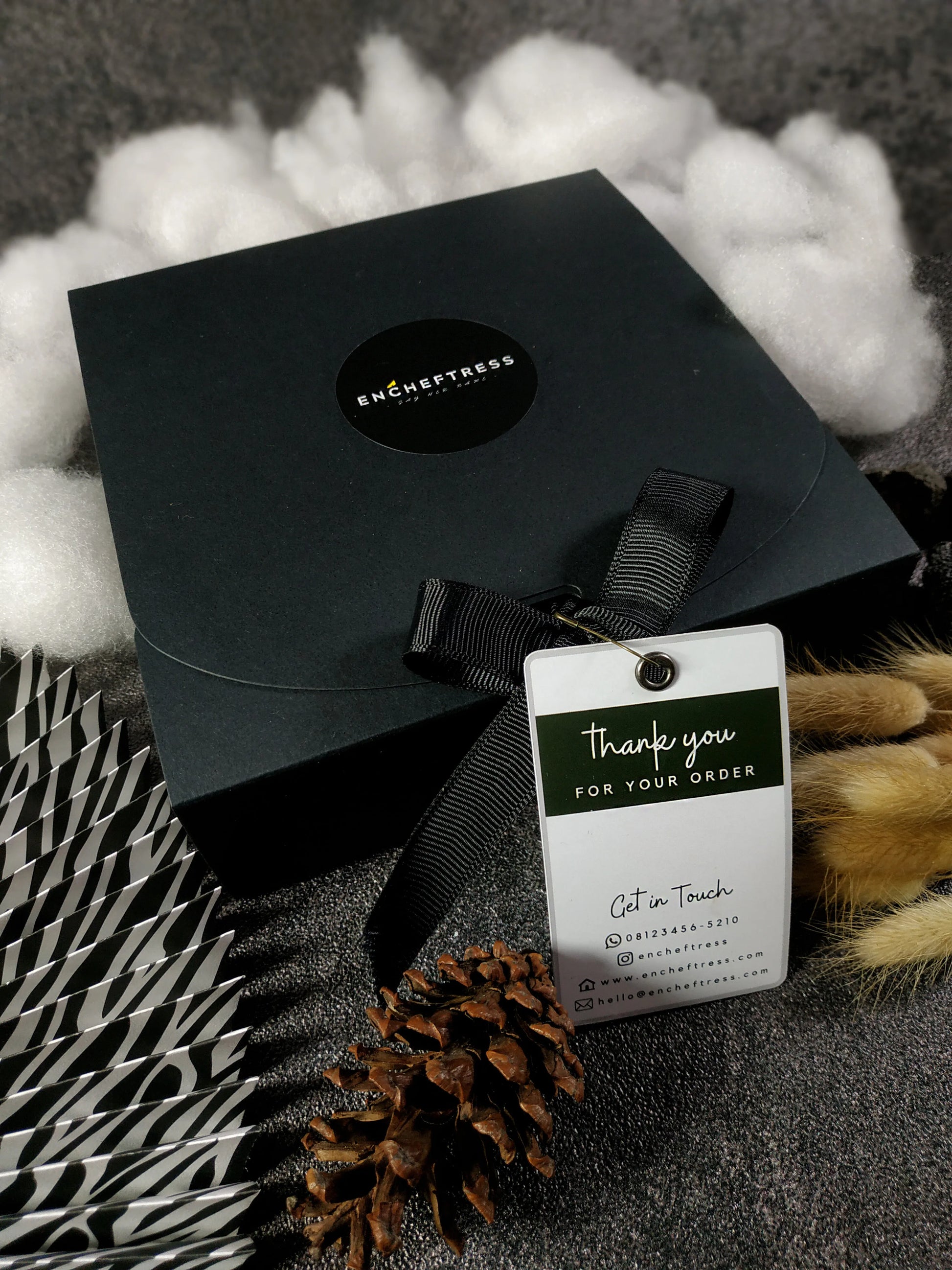 A closed black square gift box with a logo, tied with a black ribbon and a "Thank you for your order" tag, nestled among fluffy white material, a striped element, and a pinecone.