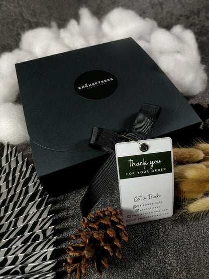 A closed black square gift box with a logo, tied with a black ribbon and a "Thank you for your order" tag, nestled among fluffy white material, a striped element, and a pinecone.