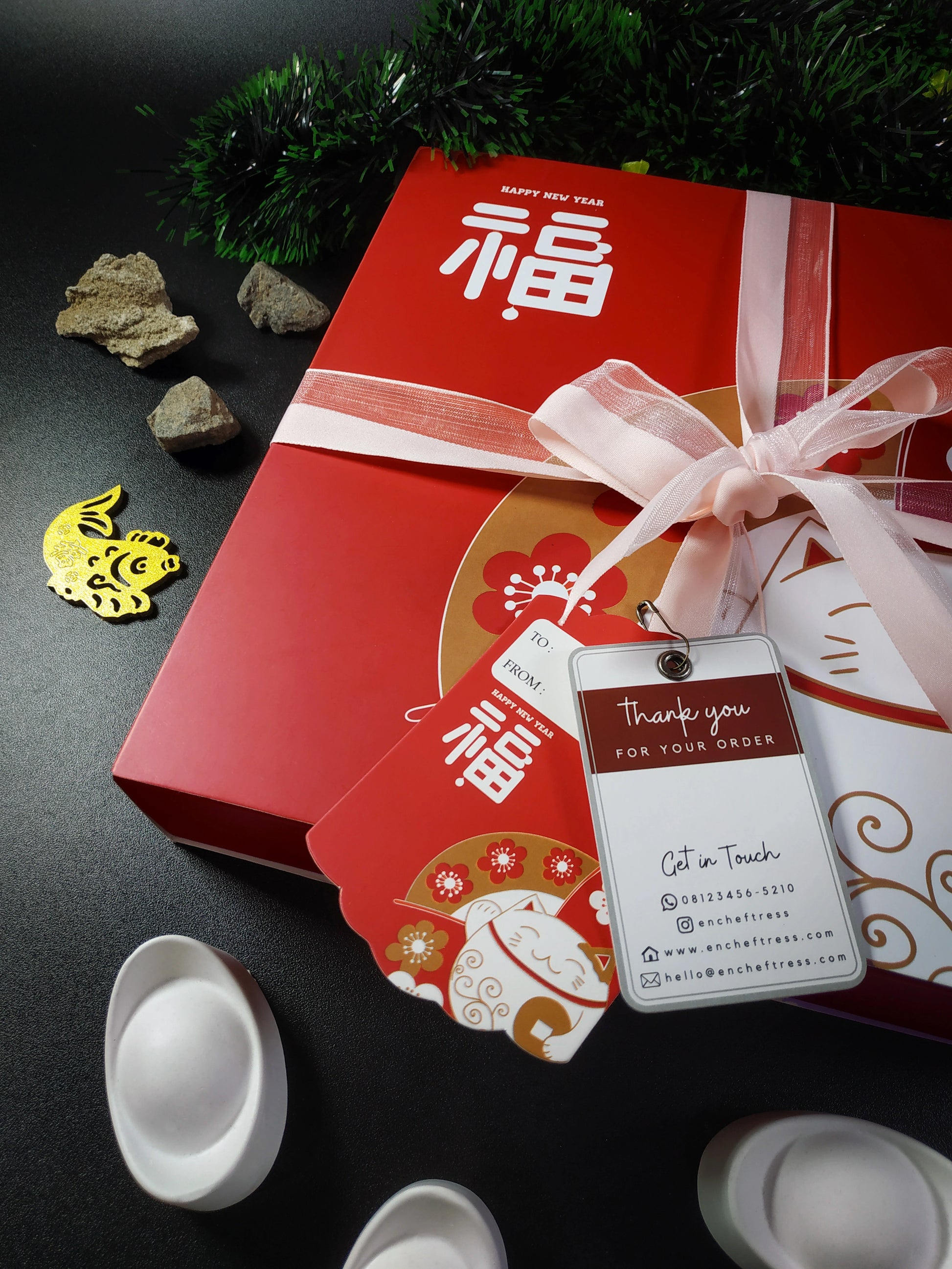 A red gift box decorated for the Lunar New Year with gold lettering and a white and pink ribbon, accompanied by a "Thank you for your order" tag, a small gold fish ornament, decorative stones, and a partial view of white packaging.