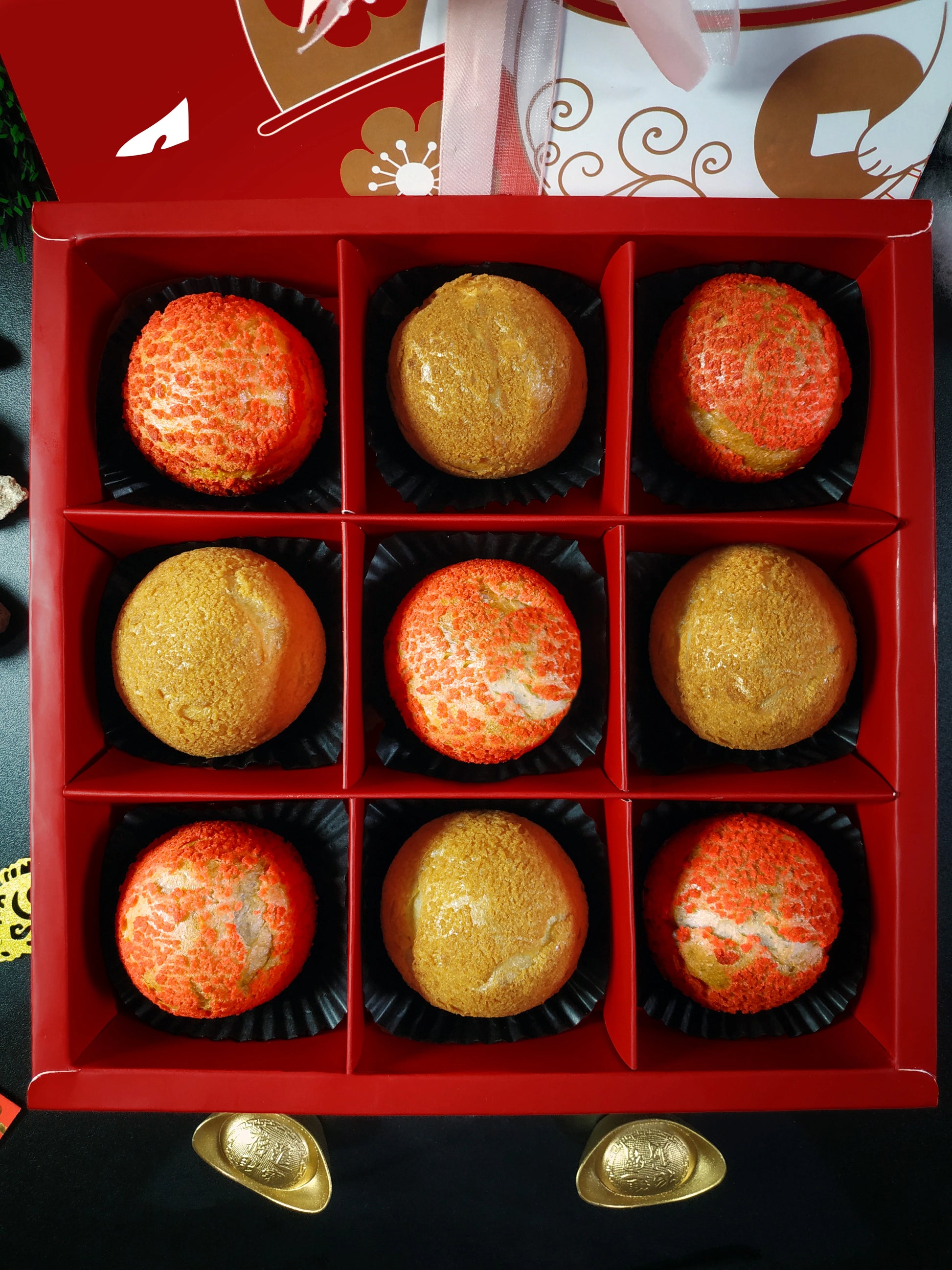 Nine colorful choux au craquelin pastries are neatly arranged in a red box with individual compartments. Gold ingot decorations are visible at the bottom of the image.