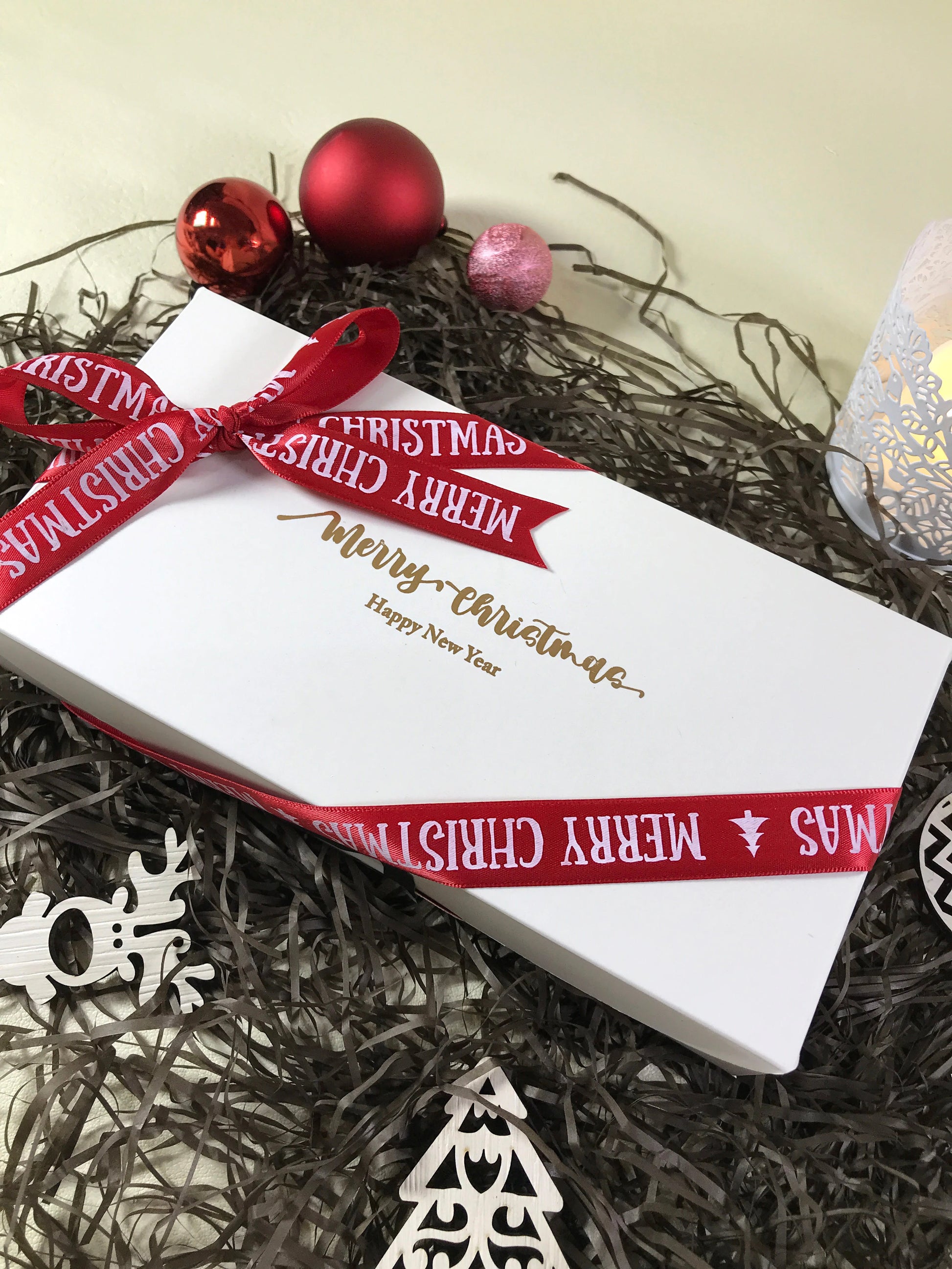 A white rectangular box with "Merry Christmas" and "Happy New Year" in gold script, tied with a red ribbon that also reads "Merry Christmas". The box is surrounded by black tinsel, red and pink Christmas ornaments, a white wooden reindeer, and a partially visible white candle holder.