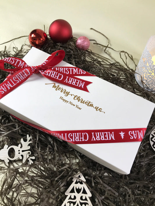 A white rectangular box with "Merry Christmas" and "Happy New Year" in gold script, tied with a red ribbon that also reads "Merry Christmas". The box is surrounded by black tinsel, red and pink Christmas ornaments, a white wooden reindeer, and a partially visible white candle holder.