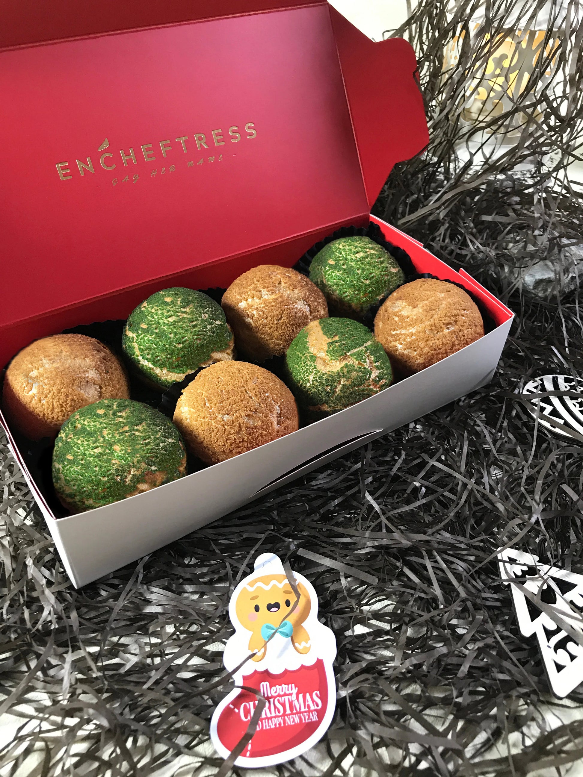 An open red and white box containing eight colorful choux au craquelin pastries, surrounded by black and silver tinsel and two white wooden Christmas ornaments. A gingerbread man cookie with a "Merry Christmas & Happy New Year" tag is visible at the bottom.