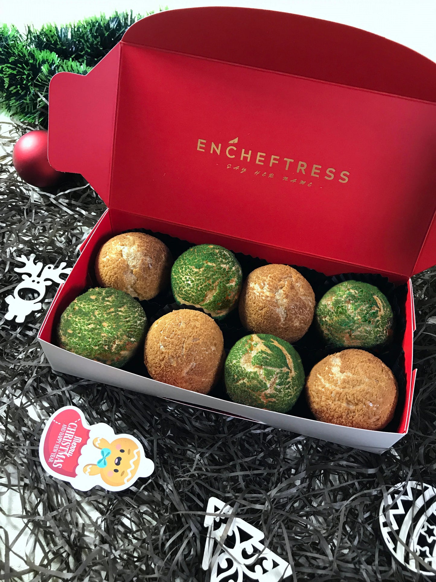 Eight colorful choux au craquelin pastries are nestled in a red and white box, surrounded by black and silver tinsel, a red Christmas ornament, and white wooden Christmas decorations. A gingerbread man cookie with a "Merry Christmas & Happy New Year" tag is also visible.