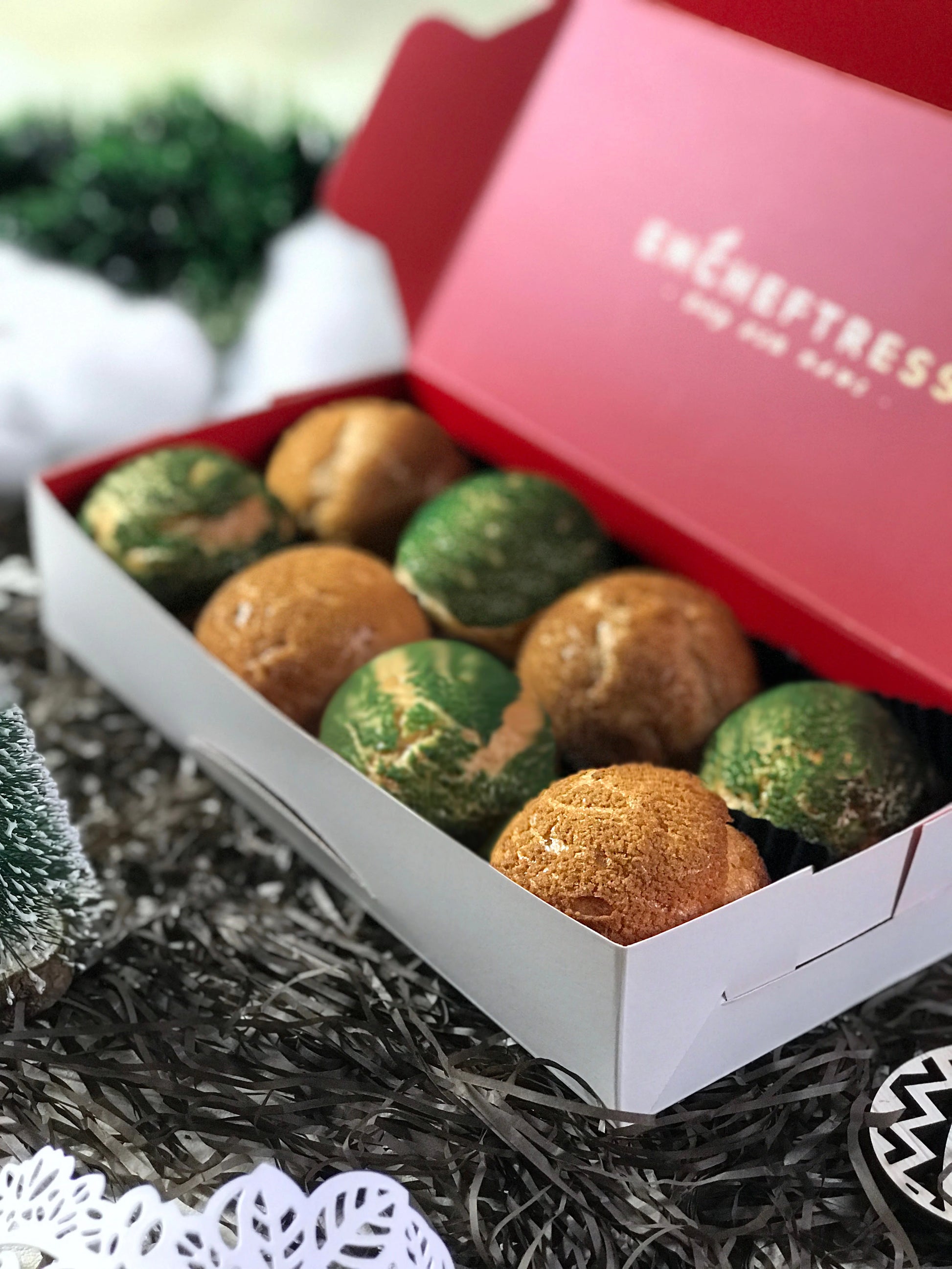 Eight colorful choux au craquelin pastries are arranged in a red and white box, surrounded by black and silver tinsel, a small Christmas tree, and white snowflake decorations.