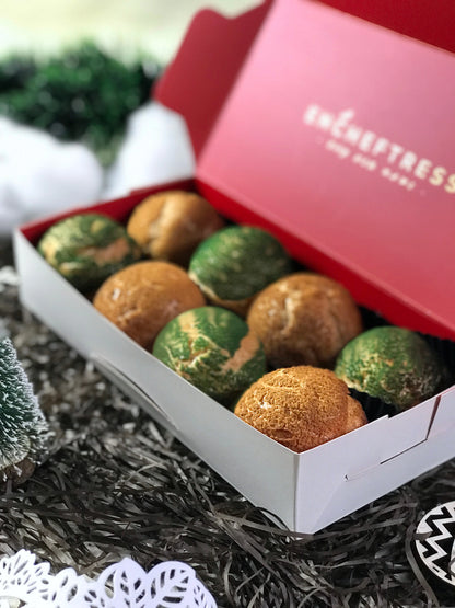 Eight colorful choux au craquelin pastries are arranged in a red and white box, surrounded by black and silver tinsel, a small Christmas tree, and white snowflake decorations.