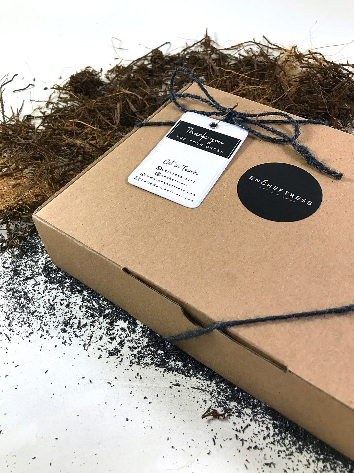 A closed brown cardboard box with a black circular logo and a "Thank you for your order" tag tied with a dark string, surrounded by scattered moss-like material and dark particles on a light surface.