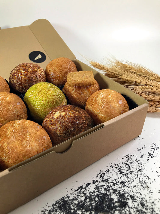 An open cardboard box reveals twelve colorful choux au craquelin pastries, with wheat stalks and scattered dark tea leaves on a white surface beside it.