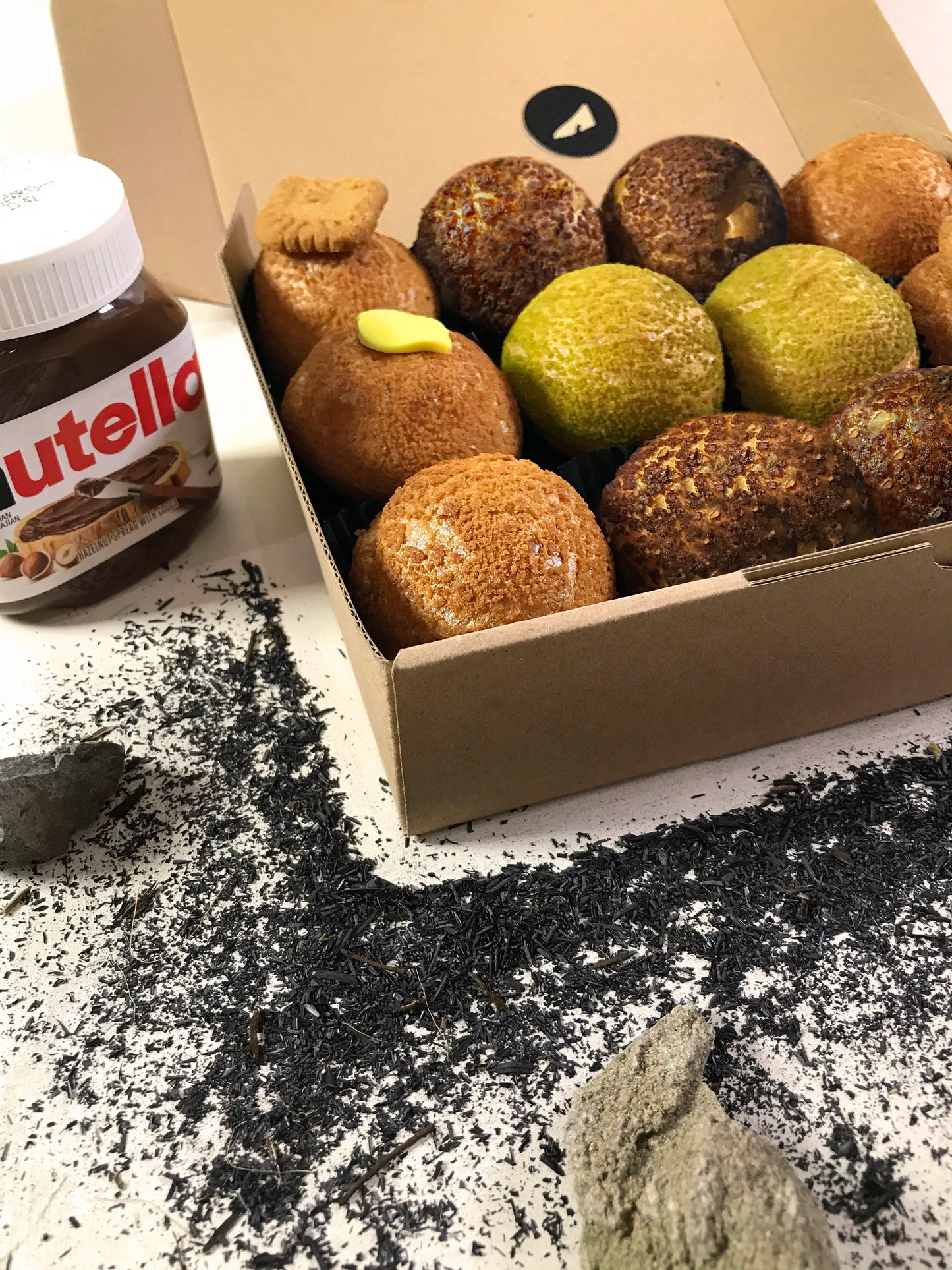 An open cardboard box containing twelve colorful choux au craquelin pastries sits on a white surface scattered with dark tea leaves and a few light-colored stones. A jar of Nutella is partially visible in the upper left corner.