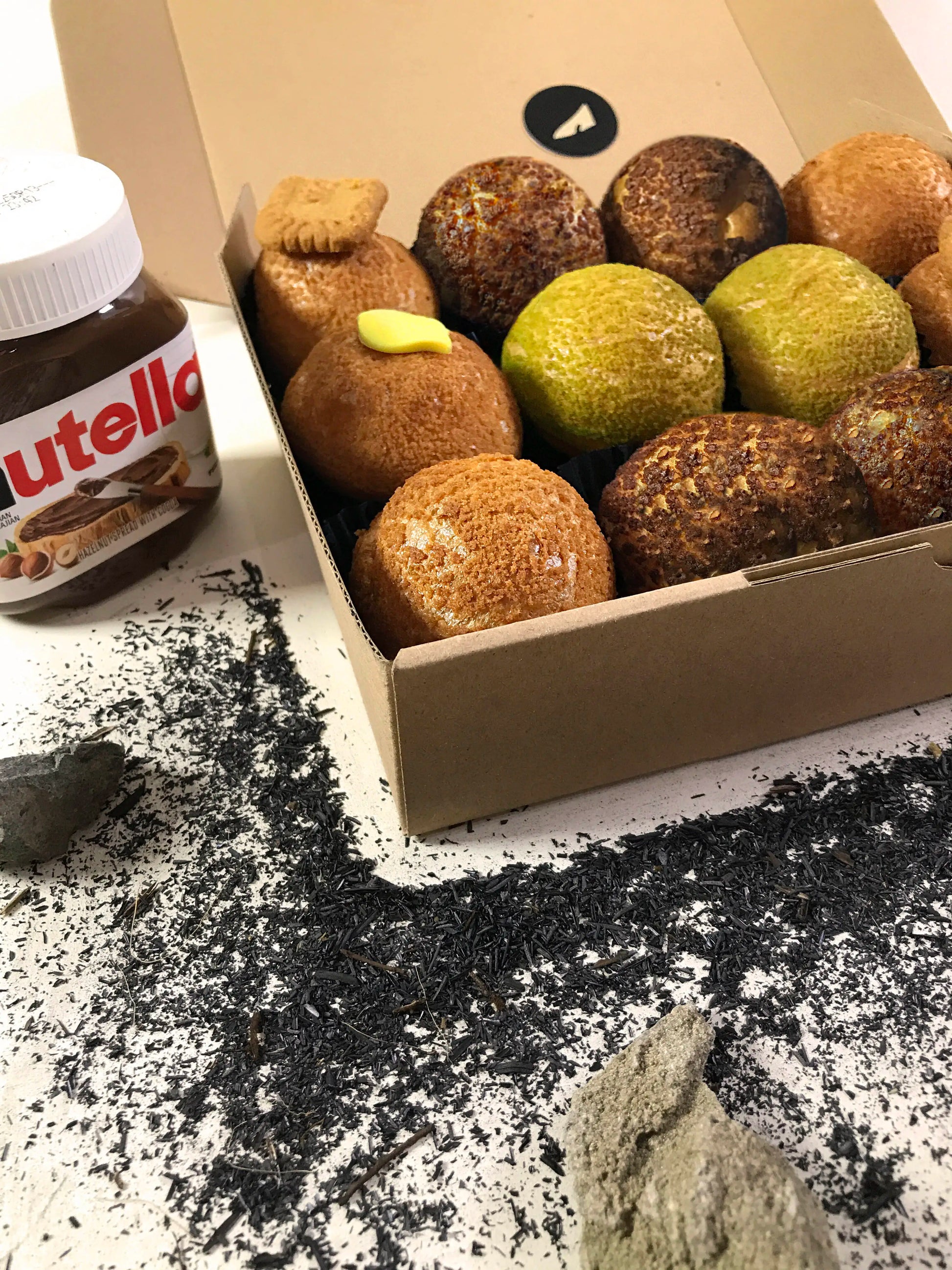 An open cardboard box containing twelve colorful choux au craquelin pastries sits on a white surface scattered with dark tea leaves and a few light-colored stones. A jar of Nutella is partially visible in the upper left corner.