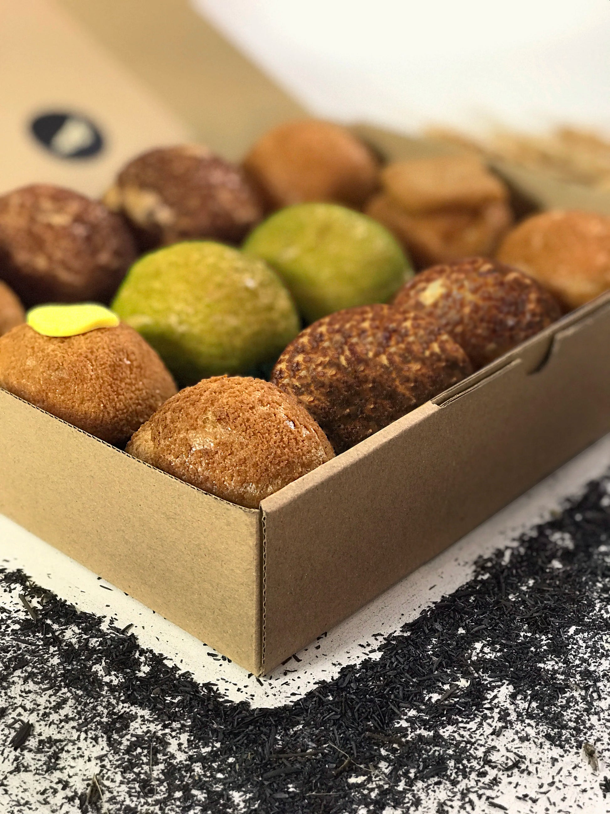 A close-up shot shows twelve colorful choux au craquelin pastries nestled inside an open cardboard box, with scattered dark tea leaves on the white surface below.