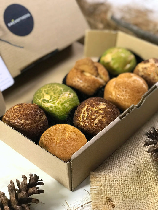 Eight colorful choux au craquelin pastries arranged in a cardboard box, with pinecones and burlap fabric visible around the box.