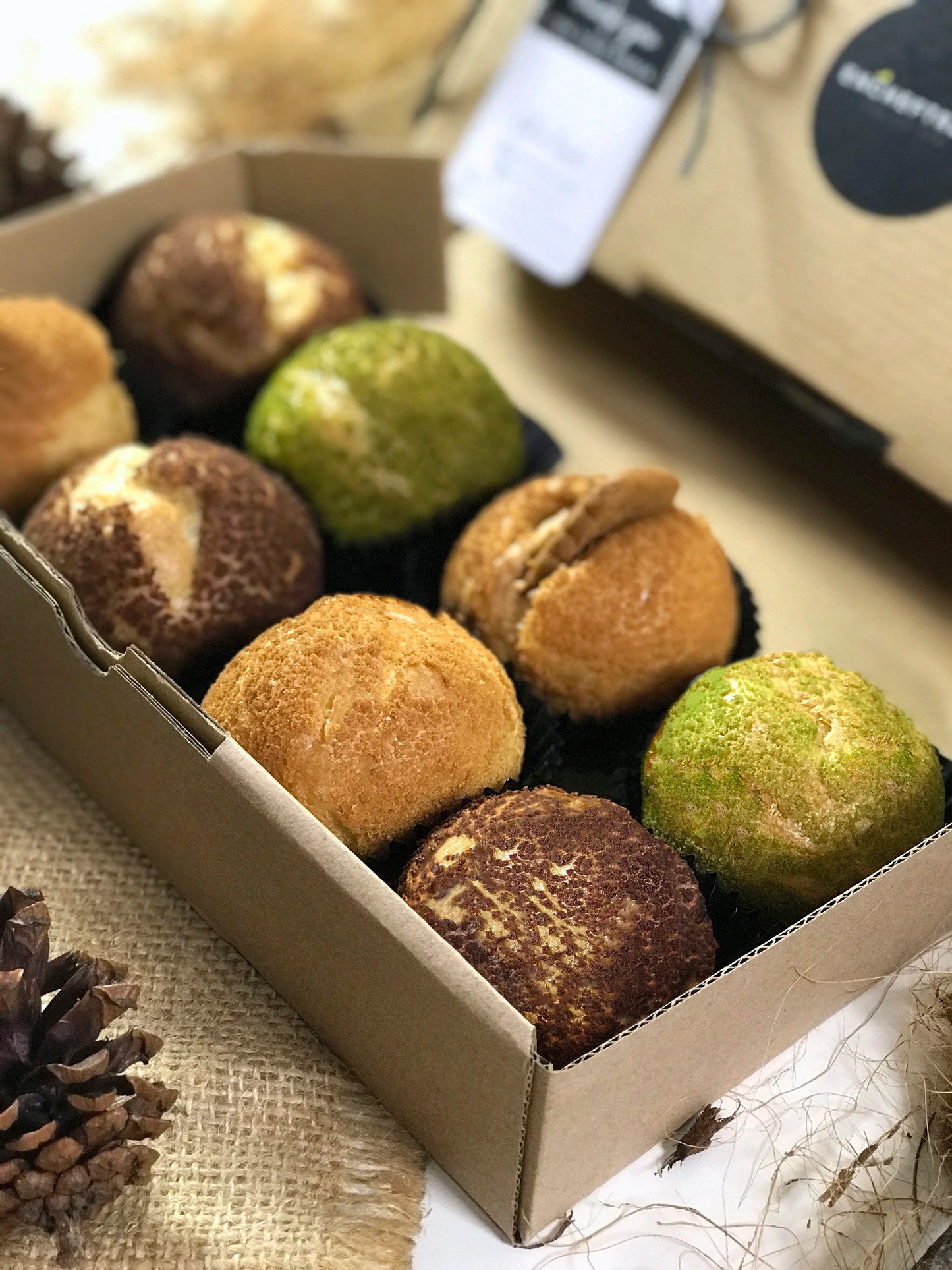 A box of eight colorful choux au craquelin pastries on a burlap surface with scattered pinecones.