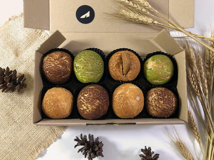 An open cardboard box containing eight colorful choux au craquelin pastries, surrounded by wheat stalks and pinecones on a white surface.