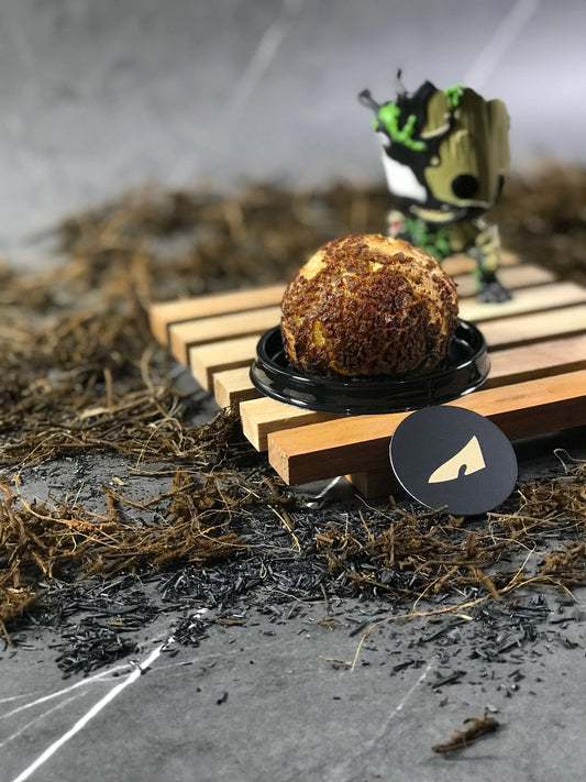 A single choux au craquelin pastry on a small wooden pallet with a Groot figurine, against a textured gray background with scattered moss-like material.