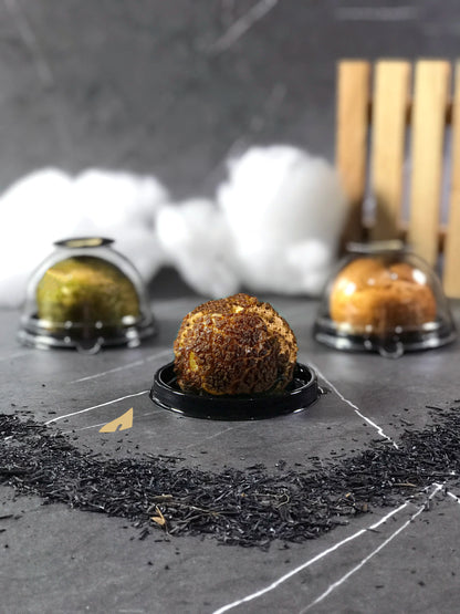 Three individually packaged choux au craquelin pastries on a gray surface with scattered dark tea leaves, against a blurred background of white cotton and a wooden structure.