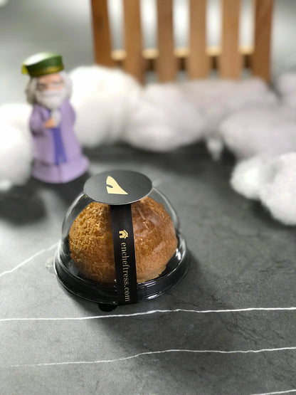 A single choux au craquelin pastry in a clear plastic container with a black base and branded sticker, with a Dumbledore figurine and fluffy white material in the blurred background.