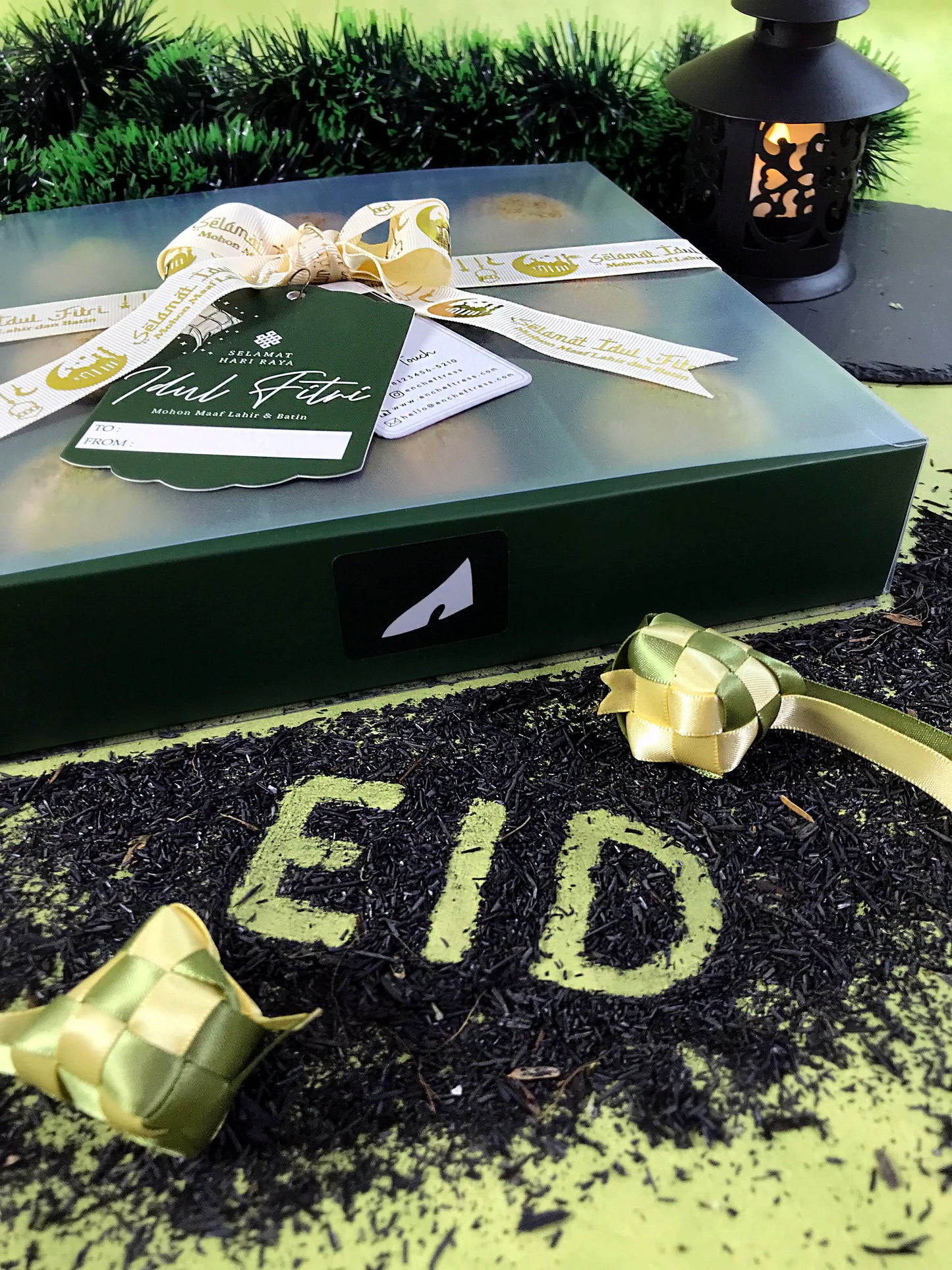 A green gift box with a white and gold ribbon and a 'Selamat Hari Raya Idul Fitri' tag, placed on a surface covered in dark tea leaves that spell out "EID" in leaf green. Two ketupat decorations and a lantern are also visible, suggesting a Ramadan or Eid theme.