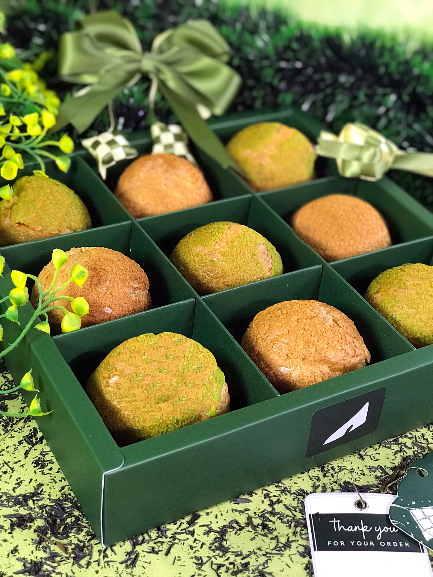Nine colorful choux au craquelin pastries are arranged in a green box with individual compartments, decorated with a green bow and a 'Thank you for your order' tag, against a backdrop of green foliage and scattered dark particles.