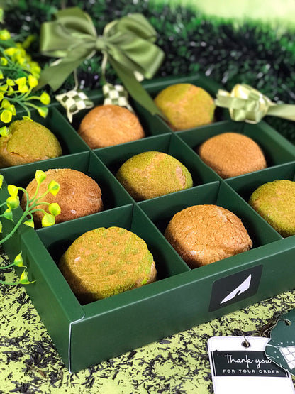 Nine colorful choux au craquelin pastries are arranged in a green box with individual compartments, decorated with a green bow and a 'Thank you for your order' tag, against a backdrop of green foliage and scattered dark particles.