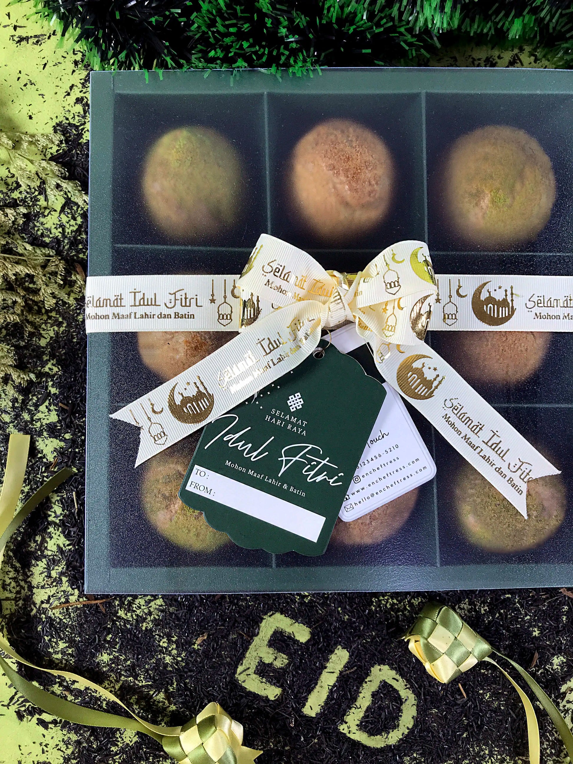 A green box containing nine choux au craquelin pastries is tied with a white and gold ribbon featuring "Selamat Idul Fitri" text, along with a matching tag. The box sits on a surface covered in dark tea leaves that spell out "EID" in leaf green, with ketupat decorations and green tinsel nearby, suggesting an Eid al-Fitr celebration.