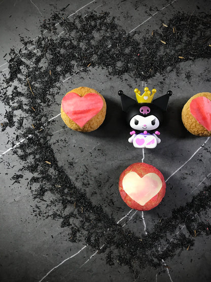 Three heart-shaped choux au craquelin pastries and a Kuromi figurine with a gold crown are arranged on a dark, textured surface scattered with black tea leaves.