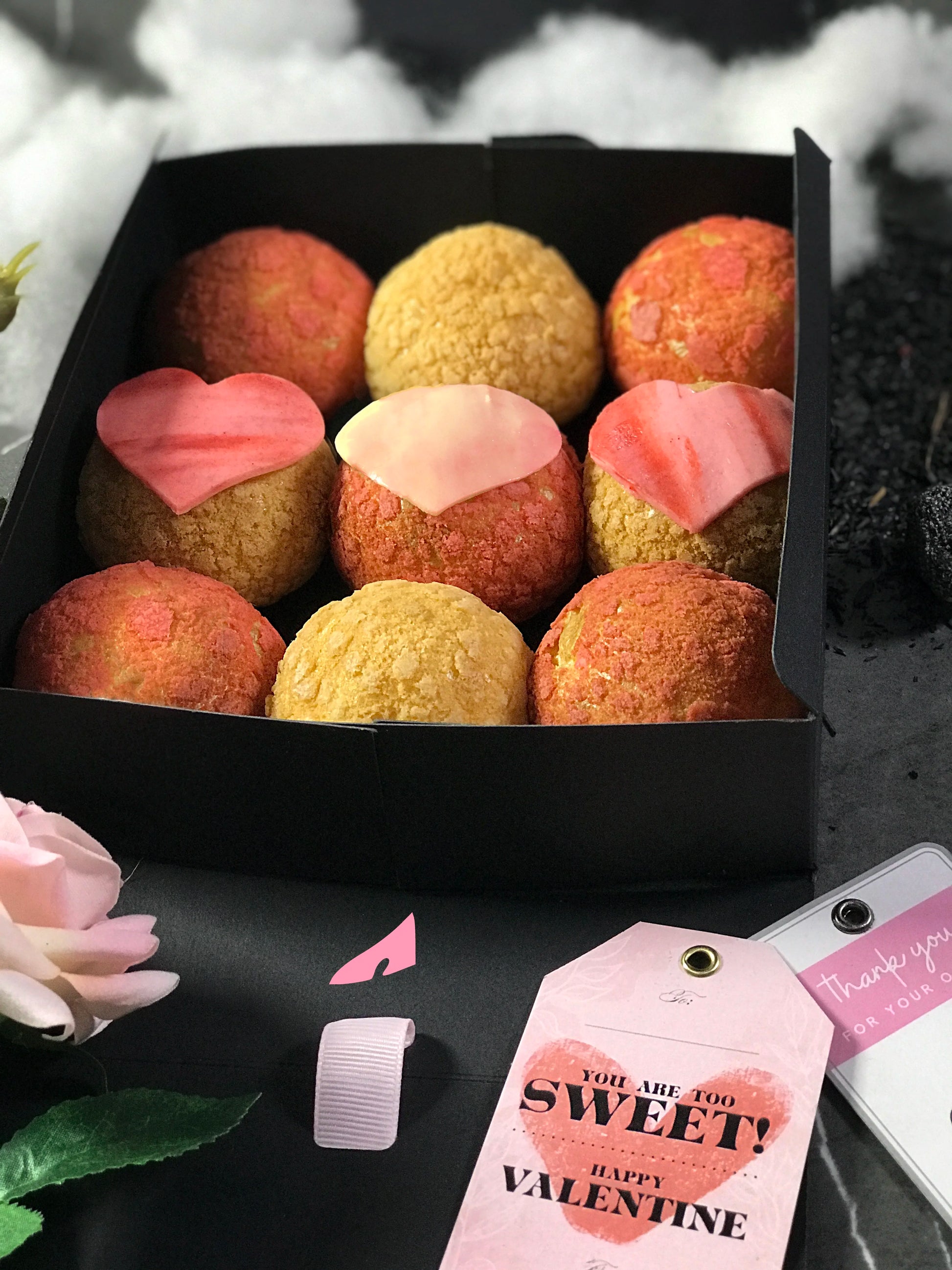 Nine colorful choux au craquelin pastries, three with heart-shaped tops, are arranged in a black box with a Valentine's Day tag that reads "YOU ARE TOO SWEET! HAPPY VALENTINE". The box is surrounded by pink and white artificial roses and scattered dark particles.