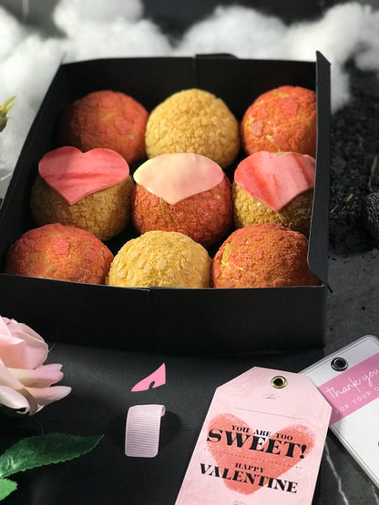 Nine colorful choux au craquelin pastries, three with heart-shaped tops, are arranged in a black box with a Valentine's Day tag that reads "YOU ARE TOO SWEET! HAPPY VALENTINE". The box is surrounded by pink and white artificial roses and scattered dark particles.