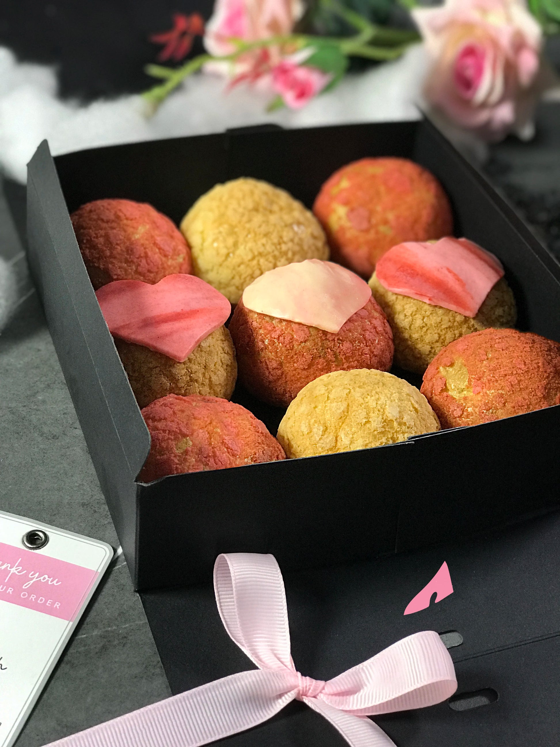 Nine colorful choux au craquelin pastries, three with pink heart-shaped tops, are arranged in a black box. A light pink ribbon and a "Thank you for your order" tag are near the box, with blurred pink and white flowers in the background.