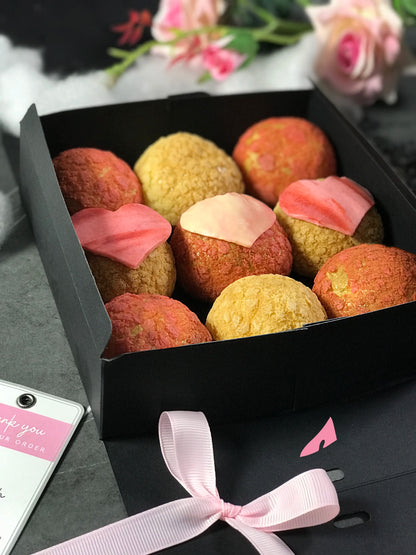Nine colorful choux au craquelin pastries, three with pink heart-shaped tops, are arranged in a black box. A light pink ribbon and a "Thank you for your order" tag are near the box, with blurred pink and white flowers in the background.