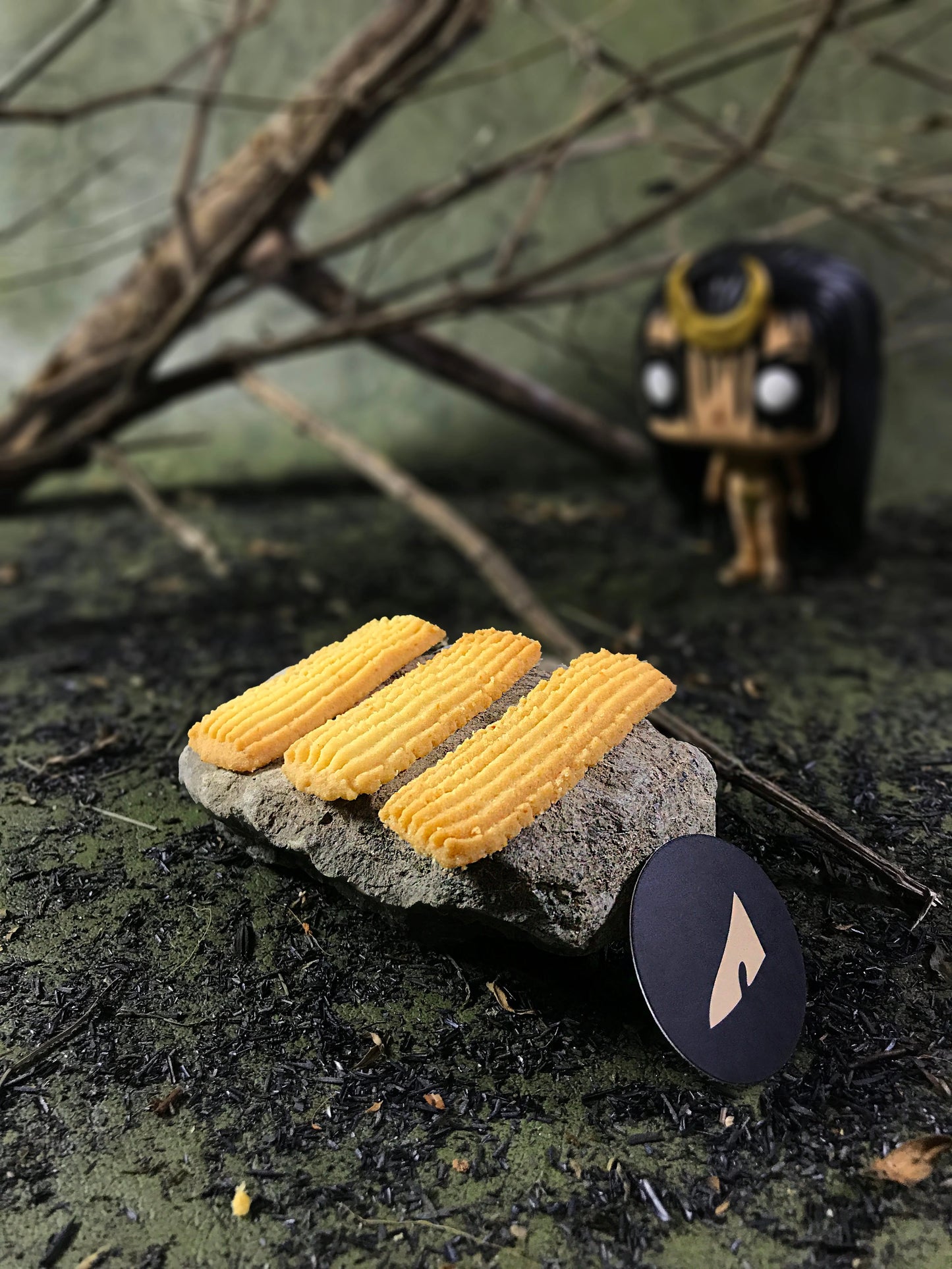 Three rectangular butter cookies with ridged tops rest on a gray stone, with a black circular tag bearing a stylized light gold shape. The setting includes blurred branches and a small, stylized enchantress figurine in the background, all on a textured, dark surface.