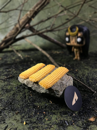 Three rectangular butter cookies with ridged tops rest on a gray stone, with a black circular tag bearing a stylized light gold shape. The setting includes blurred branches and a small, stylized enchantress figurine in the background, all on a textured, dark surface.