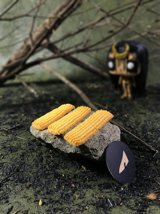 Three rectangular butter cookies with ridged tops rest on a gray stone, with a black circular tag bearing a stylized light gold shape. The setting includes blurred branches and a small, stylized enchantress figurine in the background, all on a textured, dark surface.