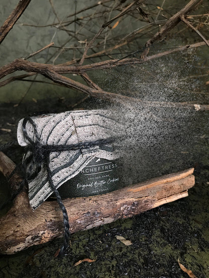 A jar labeled "Original Butter Cookies" with a black and white patterned cloth tied around the lid with black string sits on a piece of driftwood. The jar is slowly fading into tiny particles. The background includes fuzzy branches and mossy ground.