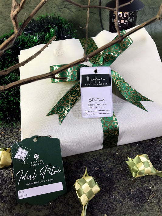 A white gift bag tied with a green and gold patterned ribbon and a "Thank you for your order" tag rests on a dark surface scattered with green tinsel and ketupat decorations. A 'Selamat Hari Raya Idul Fitri' tag and a blurred lantern are also visible.